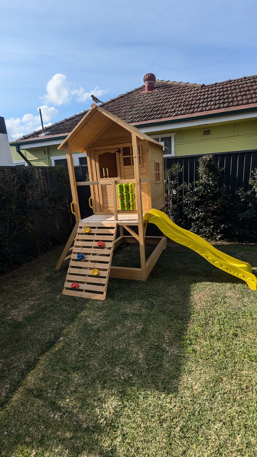 Timber cubby house with a climbing wall and yellow slide, assembled on a back lawn